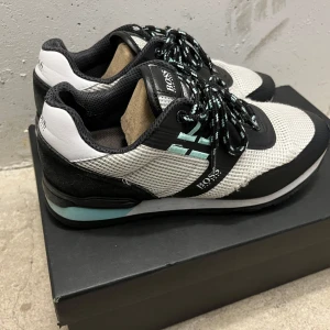 Boss skor - Snygga sneakers från BOSS med svart och vit mesh, svarta och turkosa detaljer samt logga på sidan och hälen. Skorna har snörning och en robust sula med svart och turkos rand. Perfekta för dig som gillar sportig och stilren look.