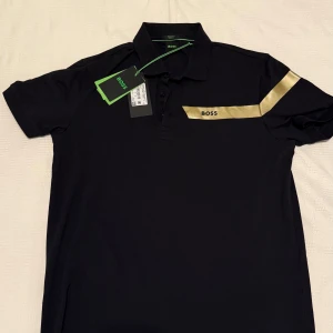 BOSS - Guld / Svart pikétröja (M) - Svart pikétröja från BOSS.  Snygg guldfärgad rand över bröstet och ena ärmen.                                     NYSKICK / OANVÄND - STORLEK : ( M )  ETIKETTER KVAR. 