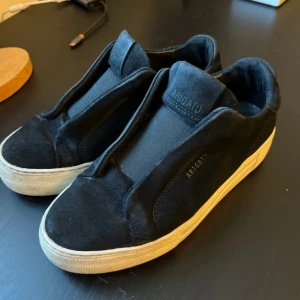 Svarta sneakers från Axel Arigato - Stilrena svarta sneakers från Axel Arigato i mocka med vit platt sula. Skorna har minimalistisk design utan snörning och diskret logga på sidan och plösen. Perfekta för dig som gillar en clean look och vill ha något som sticker ut på ett subtilt sätt ä. Köpa på vinted och säljs pga för stor storlek. Orginalpris: 2700 kr