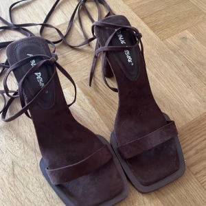 Bruna mockasandaler med klack från Public Desire - Snygga mörkbruna sandaler i mocka med fyrkantig tå och höga klackar. Skorna har långa remmar som snöras runt benet för en trendig look. Perfekta för dig som vill sticka ut med en stilren och modern vibe.