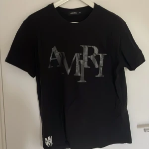 Svart AMIRI t-shirt med stor logga - Svart t-shirt från AMIRI med stor, grå logotyp framtill och mindre vit logga nere vid fållen. Klassisk passform och rund halsringning. Tillverkad i mjuk bomull, perfekt för en clean och stilren look.