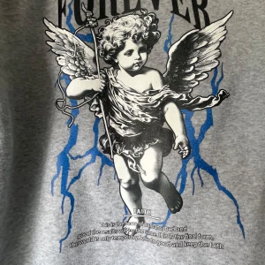 Grå hoodie med Forever-tryck och ängel - Säljer en grå hoodie med tryck av texten 'FOREVER' och en stor grafisk ängel på framsidan. Hoodien har huva med snörning och en stor magficka. Materialet är mjukt och skönt, perfekt för chill dagar. Snygga blå detaljer i trycket ger extra edge.