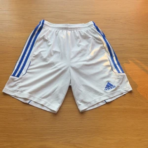 Vita Adidas shorts med blå ränder - Snygga vita Adidas shorts med blå ränder längs sidorna och broderad Adidas-logga framtill. Tillverkade i lätt och ventilerande polyester med elastisk midja och snörning. Perfekta för fotboll eller träning, klassisk sportig look.