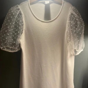 Vit topp med puffärmar från Lindex - Supersöt vit topp från Lindex med transparenta puffärmar i mesh och små prickar. Rund halsringning och rak passform. Perfekt för dig som vill ha en basic topp med en twist och extra volym på ärmarna. Små håll på ena armen💗