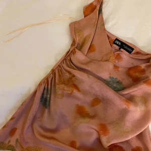 Rosa blommig linnetopp från Zara - Säljer en superfin rosa linnetopp från Zara med blommigt mönster i orange, grönt och gult. Toppen har tunna axelband, asymmetrisk nederkant och lätt draperad front. Materialet är glansigt och känns lyxigt✨