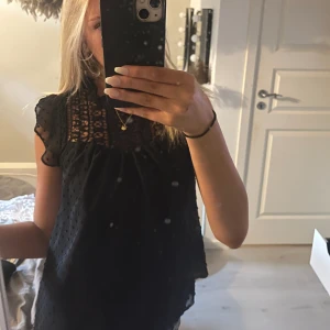 Svart blus med spets - Supersöt svart blus med transparenta partier och spets upptill. Prickig struktur i tyget och volangiga ärmar ger en romantisk vibe. Perfekt att styla med jeans eller kjol för en trendig look.