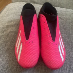 Rosa Adidas fotbollsskor utan snörning - Snygga fotbollsskor från Adidas i starkt rosa med svarta och vita detaljer. Skorna har en strumpliknande passform utan snörning, tre vita ränder på sidan och mönstrad ovandel i syntetmaterial. Sulan har fasta dobbar för bra grepp på planen.