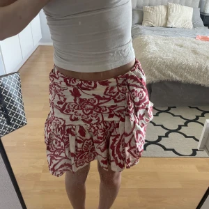 Rödvit blommig kjol från Zara - Supersöt kort kjol från Zara i storlek XS med rödblommigt mönster. Kjolen har volangkant och omlottkänsla med knyt i sidan. Perfekt för vår och sommar!