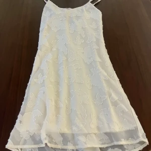 Vit kortklänning med fjärilsmönster Kappahl - Det Så fin och söt blommig klänning som är perfekt till sommaren eller till lite mer finklädda tillfällen. Bara använd 1 gång! Den är lite halvt öppen i ryggen med en tunn rosett. Original priset var 500kr💕