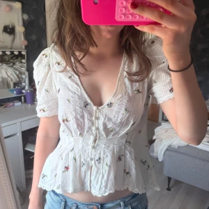 Vit blommig blus med knappar - Supersöt vit blus med broderade små blommor i olika färger. Blusen har v-ringning, puffiga korta ärmar och markerad midja. Framtill sitter klädda knappar och tyget är lätt och luftigt med fina detaljer. Perfekt för en romantisk look i sommar eller vår.