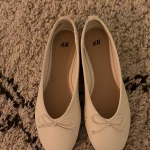 Vita ballerinaskor med rosett från H&M - Snygga vita ballerinaskor från H&M i klassisk modell med rund tå och söt rosettdetalj framtill. Skorna har en platt sula och är tillverkade i syntetmaterial, perfekta för en stilren och enkel look.