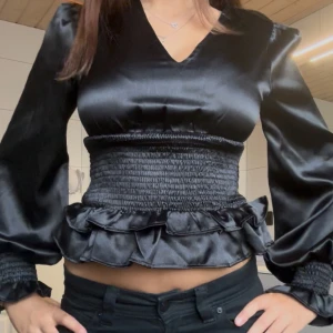 Svart satinblus med volang och smock - Supersnygg svart blus i glansigt satinmaterial med v-ringning, långa puffärmar och bred smock i midjan. Volangdetalj nedtill och smockade ärmslut ger en trendig och feminin look. Perfekt att styla med jeans eller kjol för en cool outfit.