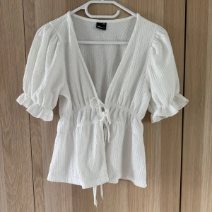 Vit blus med puffärm och knyt - Superfin vit blus från Gina Tricot med puffiga korta ärmar, djup v-ringning och knyt framtill i midjan. Blusen har en lätt textur i tyget och volangdetaljer vid ärmslut. Perfekt för en fräsch och somrig look! 