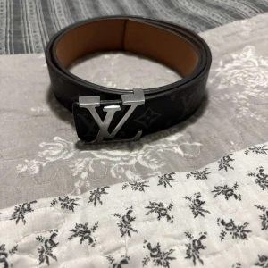Louis Vuitton svart skärp med monogram - Snyggt svart bälte från Louis Vuitton med klassiskt monogrammönster och stor LV-logga i silver på spännet. Bältet är gjort i skinn och har en stilren, exklusiv look som lyfter vilken outfit som helst.