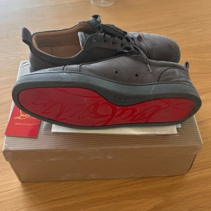 Grå mockasneakers från Christian Louboutin - Snygga grå sneakers i mocka från Christian Louboutin med ikonisk röd sula och svarta snören. Skorna har rund tå, platt sula och diskret branding på sulan. Perfekta för dig som vill sticka ut med en lyxig touch i vardagen.
