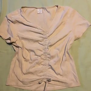 Beige omlottopp med snörning från Lindex - Snygg beige topp från Lindex med diskreta ränder och v-ringning. Toppen har korta ärmar och cool snörning framtill som ger en trendig look. Perfekt för dig som gillar en enkel men ändå unik stil.