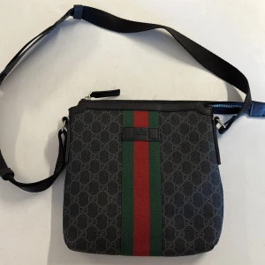 Gucci axelväska med GG-mönster - Snygg axelväska från Gucci med klassiskt GG-mönster i grått och svart. Väskan har bred grön och röd rand framtill, justerbar axelrem och dragkedja upptill. Tillverkad i canvas med detaljer i skinn. Perfekt för dig som vill ha en stilren och ikonisk accessoar.