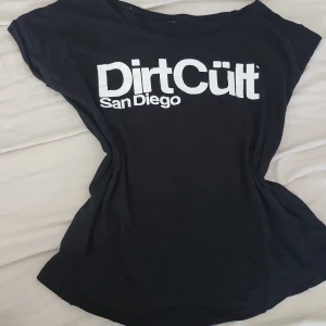 Svart Dirt Cult San Diego t-shirt - Svart t-shirt från Dirt Cult med stor vit logga och texten 'San Diego' på bröstet. Klassisk rund hals och korta ärmar. Skön och avslappnad passform, perfekt till jeans eller shorts. Materialet känns mjukt och lätt, troligtvis bomull.. Den är offshoulder och lite lång i längden. 