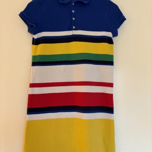 Randig pikéklänning från Polo Ralph Lauren, storlek XXS/ 12-14 år - Pikéklänning från Polo Ralph Lauren i bomull och klassiska färger; Blå krage och knappar framtill och ränder i blått, vitt, gult, grönt, rött och svart. Bra skick - en liten fläck bak dom syns på sista bilden - därav lågt pris.