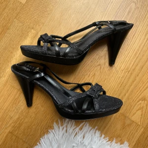 Glittriga svarta sandaler med klack - Snygga svarta sandaler från American Eagle med glittrig finish, öppen tå och hög klack. Remmarna är tunna och pryds av en söt rosett framtill. Justerbar rem runt ankeln för bästa passform.