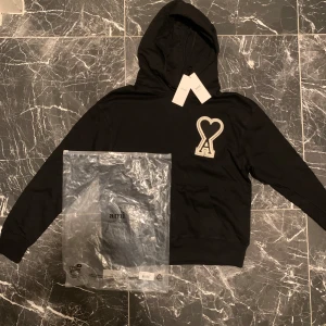 Svart hoodie från AMI Paris med logga - Svart hoodie från AMI Paris med stor vit broderad hjärtlogga och bokstaven A på bröstet. Klassisk känguruficka, huva med snörning och diskret AMI-broderi i nacken. Skön passform och perfekt för chill dagar.