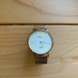 Guldig klocka Lorenzo Milano meshband - Stilren klocka från Lorenzo Milano med vit urtavla, guldiga detaljer och ett elegant meshband i metall. Minimalistisk design med tunna index och liten italiensk flagga på urtavlan. Perfekt accessoar för dig som gillar klassisk stil. Behöver nytt batteri 