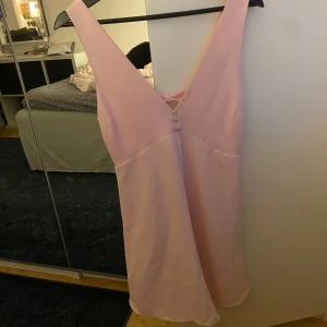Ljusrosa kort  klänning från Zara - Supersöt ljusrosa kort klänning från Zara med djup v-ringning både fram och bak, dekorerad med små knappar framtill och en rosett i ryggen. Klänningen är ärmlös och har en figursydd men lätt utsvängd passform. Perfekt för middagar o fester nu i sommar!!! Aldrig använd!