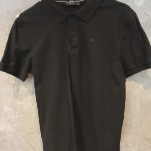 Svart pikétröja från J.Lindeberg - Klassisk svart pikétröja från J.Lindeberg med korta ärmar och krage. Tröjan har diskret logga på bröstet och är tillverkad i bomull. Perfekt för en clean och stilren look.