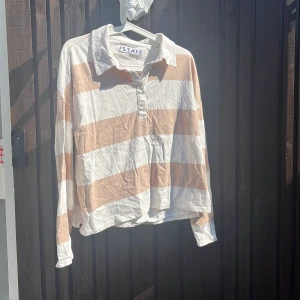 Randig pikétröja från Stay i beige och vitt - Säljer en oversized pikétröja från Stay i storlek M. Tröjan har breda beige och vita ränder, klassisk krage och knäppning med tre knappar framtill. Långärmad och tillverkad i mjuk bomull, perfekt för en chill och avslappnad stil.