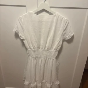 Vit omlottklänning från Dressy - Supersöt vit kortklänning från Dressy med omlott och knyt framtill, smockad midja och volangdetaljer vid ärmslut och kjolkant. Perfekt för sommarens alla tillfällen och ger en fräsch och romantisk vibe.