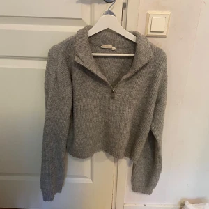 Grå stickad half zip tröja - Mysig grå stickad tröja med half zip och hög krage. Tröjan har en ribbad struktur och långa ärmar, perfekt för kyliga dagar. Enkel att matcha med jeans eller mjukisbyxor för en chill look.