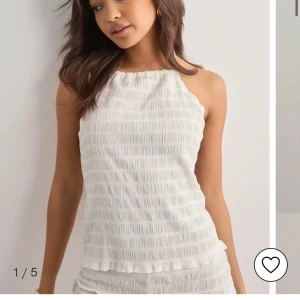 Vit ärmlös linnetopp med knyt från Nelly  - Supersöt vit linnetopp med smockad struktur och tunna knytband i nacken och ryggen. Toppen är ärmlös och har öppen rygg, perfekt för varma dagar. Lätt och luftig känsla med volangkant nertill. Enkel att styla till shorts eller kjol.