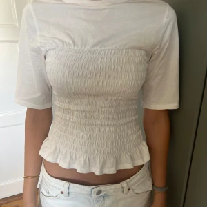 Vit smockad topp med volangkant - Supersöt vit topp med smockad midja och volangkant nertill. Modellen är croppad med korta ärmar och rund halsringning. Perfekt att styla med jeans eller kjol för en fräsch look. Gjord i mjuk bomull som känns skön mot huden.