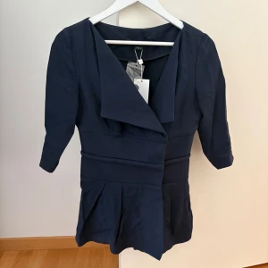 Marinblå blazer Porsche Design - Snygg marinblå blus/blazer från Porsche Design, tillverkad i Italien. Klänningen har trekvartsärm, bred krage och markerad midja med veckad kjol. Stängs med tryckknappar framtill och även knappar på insidan. Den är i strl 36, och aldrig använd med prislapp kvar! Det kan vara bra att veta att materialet inte är särskilt töjbart. 💙