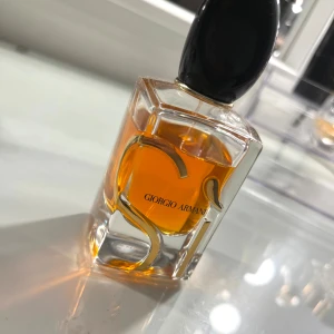 Giorgio Armani Sì Eau de Parfum - Lyxig parfymflaska från Giorgio Armani med elegant, rektangulär form och svart oval kork. Flaskan är transparent med guldiga detaljer och innehåller en djup bärnstensfärgad vätska. Perfekt för dig som gillar exklusiva dofter och snygg design.
