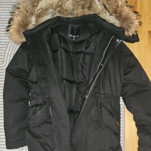 Svart dunparkas med päls från Cedrico - Säljer en svart dunparkas från Cedrico med fluffig päls runt huvan och quiltad insida. Jackan har dragkedja, praktiska fickor och snygga detaljer i lammnappa. Yttermaterialet är bomull och polyester, fodret är dun och pälsen är tvättbjörn/kanin. Perfekt för kalla dagar.