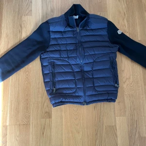 Mörkblå pufferjacka från Moncler - Snygg mörkblå pufferjacka från Moncler med stickade ärmar och quiltad front. Jackan har två dragkedjeförsedda fickor, ribbad krage och Moncler-logga på ärmen. Perfekt för dig som vill ha en stilren och exklusiv look under höst och vinter.