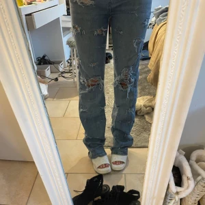 Blå ripped straight jeans - Säljer ett par blå jeans med rejält slitna och trasiga detaljer framtill och på benen. Modellen är straight fit med normal passform och klassisk femficksdesign. Jeansen har hög midja och är gjorda i jeansmaterial. Perfekt för dig som gillar en edgy och avslappnad stil.