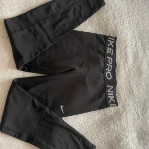 Nike Pro svarta leggings - Svarta Nike Pro leggings med bred elastisk midja där det står NIKE PRO i vitt. Snyggt swoosh-tryck på benet. Stretchigt syntetmaterial som sitter tight och är perfekt för träning. Sportig och clean look som funkar till gymmet eller löprundan.