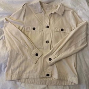 Beige overshirt med knappar - Snygg beige overshirt med klassisk krage och svarta knappar framtill. Jackan har två bröstfickor med lock och knapp, samt knappar vid ärmslut och axlar. Tillverkad i bomull, perfekt för lager-på-lager och chill stil. Sitter mer som en M