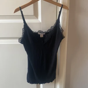Svart ribbad linnetopp med spets från new yokerAmisu - Svart linnetopp från new yoker i storlek XS med ribbad struktur och snygga spetsdetaljer längs v-ringad halsringning och axelband. Tunn, mjuk och stretchig, perfekt att styla med jeans eller kjol. Justerbara axelband för bästa passform.