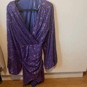 Lila glittrig omlottklänning  - Säljer en lila kortklänning med glittriga paljetter över hela klänningen. Modellen har omlott-design, v-ringad hals och långa puffärmar. Klänningen har en snygg drapering framtill och är perfekt för dig som vill sticka ut med färg och glitter. Pris kan diskuteras 