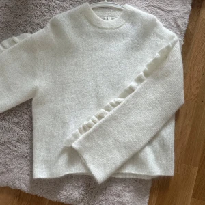 Vit stickad tröja med volangdetalj - Superfin vit stickad tröja med volangdetaljer längs ärmarna. Tröjan har en rund halsringning och långärmade, lite loose passform. Perfekt för dig som gillar mjuka och fluffiga material med en extra touch på ärmarna.