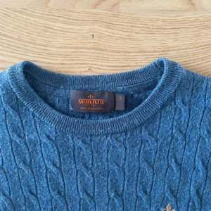 Blå kabelstickad tröja Morris - Snygg blå kabelstickad tröja från Morris i 100% merinoull. Tröjan har rund hals, lång ärm och ett litet broderat emblem på bröstet. Perfekt för dig som gillar klassisk stil med en modern touch. Passformen är normal och materialet är mjukt och värmande. Den är nästan ny den är knappt använd. Hör av er vid minsta fundering