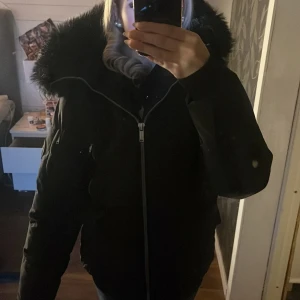 Svart pufferjacka med fuskpäls - Säljer en svart pufferjacka från Space X Demand med stor huva och fluffig fuskpäls. Jackan har dragkedja framtill, flera fickor med både dragkedja och lock, samt quiltad design. Perfekt för kalla dagar och riktigt snygg till jeans helt ny nästan.