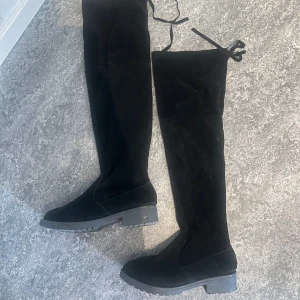 Svarta overknee mockastövlar - Säljer ett par svarta overknee stövlar i mjuk mocka med platt sula och rund tå. Stövlarna har snörning upptill som detalj och grov yttersula för extra grepp. Perfekta för att lyfta din outfit under kyliga dagar.