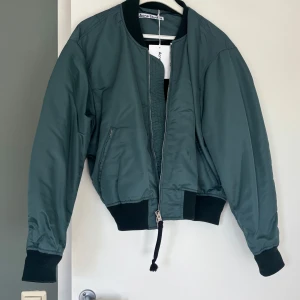Grön bomberjacka från Acne Studios - Snygg grön bomberjacka från Acne Studios med klassisk siluett, ribbade muddar och midja. Jackan har dragkedja med svart snörningsdetalj och två fickor framtill. 