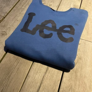 Blå sweatshirt från Lee med tryck - Snygg blå sweatshirt från Lee med stort svart logotryck på bröstet. Tröjan har rund hals, långa ärmar och ribbade muddar vid ärmslut och nederkant. Perfekt för dig som gillar enkel och stilren streetwear med tydlig branding. Axelbredd: 43cm Längd: 64cm