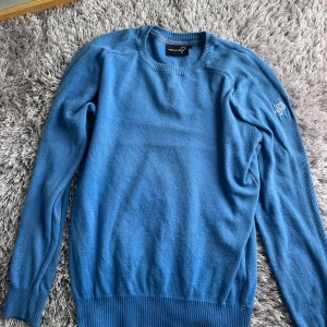 Blå stickad tröja från Pelle P - Snygg blå stickad tröja från Pelle P med rund hals och ribbade muddar vid ärmslut och nederkant. Tröjan har lång ärm och en diskret logga broderad på ena ärmen. 