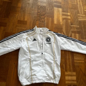 Chelsea FC vit vindjacka Adidas  - Chelsea FC vindjacka från Adidas i vit med svarta och guldiga detaljer. Jackan har dragkedja framtill, svarta ränder längs ärmarna och broderade loggor på bröstet. Lätt och sportig design, perfekt för fotbollsfans. Snyggt tryck med Chelsea FC på ryggen.